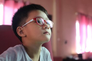 Gözlüklü bir çocuk içeride oturur, perdeye çok odaklanır. Yumuşak ışıklandırma, onun ilginç ifadesini gösteren rahat bir atmosfer yaratıyor..