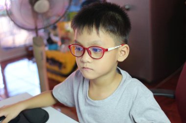 Kırmızı gözlüklü genç bir çocuk bilgisayar ekranına odaklanmış bir masada oturuyor. Ayarlar, teknoloji ve öğrenmenin vurgulandığı modern bir çalışma alanını yansıtıyor..