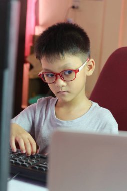 Gözlüklü odaklanmış genç bir çocuk bir masada oturuyor, dizüstü bilgisayarla meşgul oluyor. Konsantrasyonu dijital çağda öğrenmenin önemini yansıtıyor..