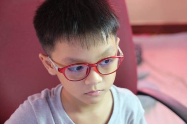 Şık kırmızı gözlük takan genç bir çocuk düşünceli bir şekilde kapalı bir yerde oturur. Onun ciddi ifadesi derin konsantrasyon ve merakı yansıtır..