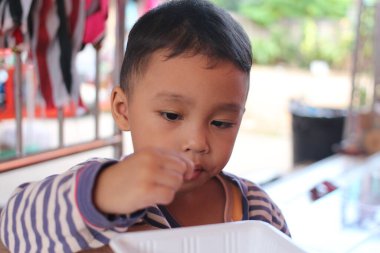 Dışarıda oturan küçük bir çocuk, atıştırmanın tadını çıkarmaya odaklanmış. Masumiyet, neşe ve gündelik bir atmosfer yakaladığı an oyuncu bir merak sergiliyor..