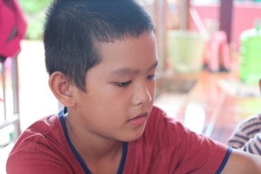 Kırmızı tişörtlü genç bir çocuk evde sessiz hareketler yaparken düşünceli bir yüz ifadesi sergiliyor. İçerideki sıcak ortam konsantrasyonunu ve masumiyetini vurguluyor..