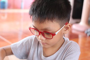 Kırmızı gözlüklü genç bir çocuk içeride oturuyor, bir aktiviteye derinden odaklanmış. Düşünceli ifadesi merakın ve öğrenmenin özünü yansıtıyor..