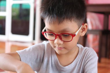 Kırmızı gözlük takan odaklanmış genç bir çocuk evde yaratıcı bir öğrenme etkinliğine katılıyor, oyuncu ortamında merak ve bağlılık gösteriyor.