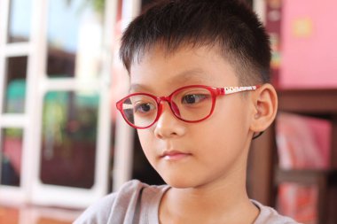 Kırmızı gözlük takan düşünceli genç bir çocuk dikkatle bakıyor, kapalı bir ortamda konsantrasyonunu gösteriyor. Eğitim ve çocukluk temaları için mükemmel.