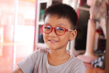 Kırmızı gözlüklü neşeli genç bir çocuk sıcak bir iç mekanda sıcacık gülümsüyor. Bu portre, gündelik bir atmosferde gençliğin neşe ve neşesini yansıtıyor..