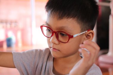 Kırmızı gözlük takan genç bir çocuk bir aktivite sırasında konsantrasyon gösteriyor. Yakın plan, onun düşünceli ifadesini yakalıyor. Merakını ve masumiyetini yansıtıyor. Parlak, neşeli bir çevrede..