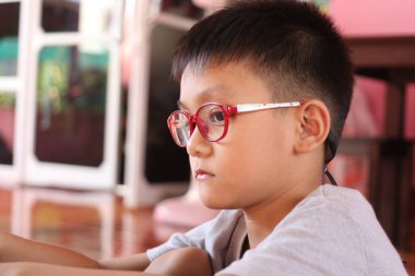 Kırmızı gözlük takan genç bir çocuk, hareketli bir iç mekanda sessizce oturur. Yüzündeki düşünceli ve meraklı ifadeyi yansıtır..