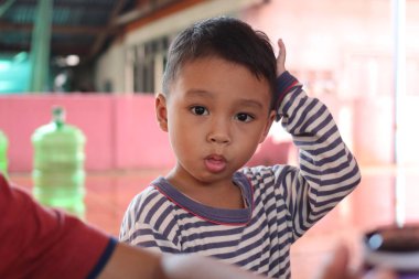 Kısa saçlı, çizgili pijama giyen genç bir çocuk ev ortamında merak gösteriyor. Onun neşeli ifadesi çocukluğun masumiyetini yakalar..