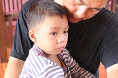 Bir babanın küçük oğlunu kucağına alıp sıcak bir ev ortamında sevgi ve bakım bağını vurguladığı dokunaklı bir an. Aile temaları için mükemmel.