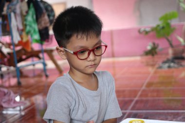 Kırmızı gözlük takan düşünceli genç bir çocuk evde oturuyor, odaklanmış ve sakin. Sıcak atmosfer, onun duygularını gelişigüzel bir ortamda ön plana çıkarıyor..