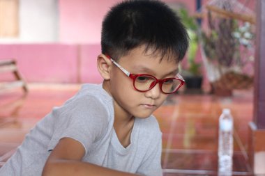 Kırmızı gözlük takan genç bir çocuk düşünceli bir şekilde kapalı bir alanda faaliyet gösteriyor. Sıcak tonlar ve doğal ışık çocukluk öğrenimi ve keşfi için ideal dostane, davetkar bir atmosfer yaratır..