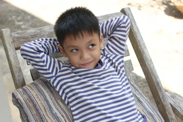 Genç bir çocuk dışarıda neşeli bir ifadeyle rahatlar. Çizgili bir gömlek giymiş, tahta bir sandalyede dinlenirken huzurlu bir anın tadını çıkarıyor..