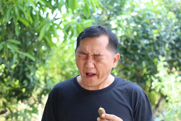 Bir adam yemyeşil bir bahçede meyve tadarken dramatik bir tepki verir. Yüzündeki ifade şaşkınlık ve hoşnutsuzluk anını yakalıyor. Taze yemek tatma sevincini gözler önüne seriyor..