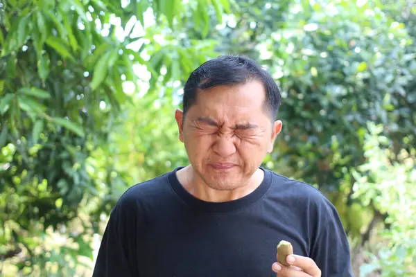 Bir adam açık hava bahçesinde alışılmadık bir yemeğin tadına bakarken güçlü bir ifade sergiliyor, gün ışığında bir şaşkınlık ve merak anı yakalıyor..