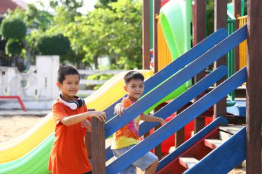 İki neşeli çocuk renkli bir oyun alanında eğlenceli aktivitelere girişiyor, güneşli bir ortamda slaytları keşfediyor ve binalara tırmanıyor..