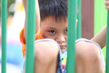 Genç bir çocuk oyun parkında oturmuş parmaklıklardan merakla bakıyor. Düşünceli ifadesi masumiyet ve keşif anlarını yakalıyor..