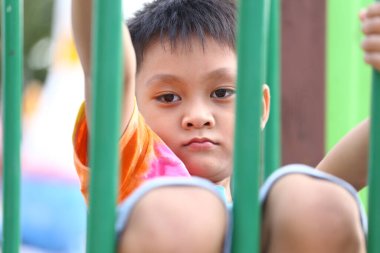 Genç bir çocuk, canlı bir tişört sergileyerek çocuk parkındaki barlarda düşünceli bir şekilde oturur. Görüntü, bir dış arkaplan üzerinde düşünme anını yakalar..