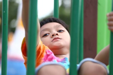 Küçük bir çocuk oyun parkı ekipmanlarına tırmanmaktan zevk alıyor, neşe ve keşif gösterisi yapıyor. Canlı renkler açık havada eğlenceli bir atmosferi vurgular..