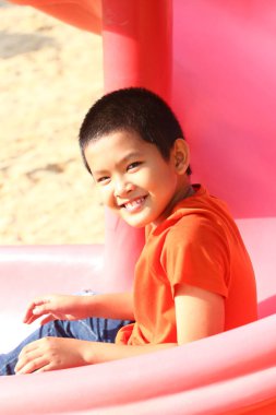 Neşeli bir çocuk renkli bir oyun parkı kaydırağında güneşli bir günün tadını çıkarıyor. Saf neşe ve çocukluk oyunlarının ve açık hava ortamının özünü sergiliyor..