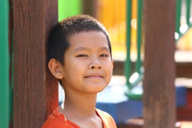 Neşeli genç bir çocuk, parlak bir oyun parkında tahta bir direğe yaslanırken gülümsüyor, neşe ve kaygısız çocukluk anlarını yakalıyor..