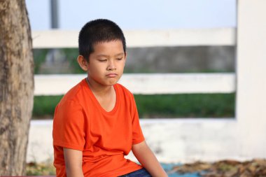 Turuncu gömlekli düşünceli bir çocuk dışarıda sakince oturur, çevresini yansıtır. Bu sakin sahne çocukluğun özünü doğal bir ortamda yakalar..