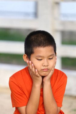Turuncu gömlekli dalgın genç bir çocuk dışarıda oturup çenesini ellerinin üzerinde dinlendiriyor. Düşünceli ifadesi derin duygular ve içgözlem taşıyor..