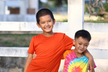 İki neşeli çocuk açık hava ortamında mutlu bir şekilde gülümsüyor, dostluk ve kaygısız anlar sergiliyorlar. Renkli kıyafetleri sahneye canlılık katıyor..
