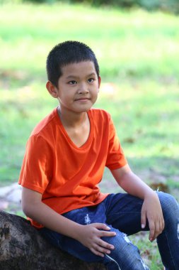 Güneşli bir açık hava ortamında bir ağaç gövdesinde gelişigüzel oturan genç bir çocuk. Parlak turuncu bir tişört giyiyor, doğada mutluluk ve rahatlık sergiliyor..