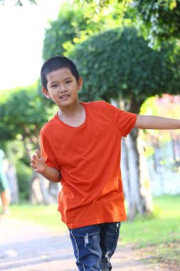 Turuncu tişörtlü neşeli genç bir çocuk neşeli bir şekilde açık hava parkında yürüyor, yemyeşil, kaygısız çocukluğun özünü yakalıyor..