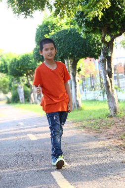 Genç bir çocuk güvenli bir şekilde ağaçlarla kaplı bir patikada yürüyor, neşe ve macerayı somutlaştıran güneşli, canlı bir açık hava ortamında yeşilliklerle dolu.