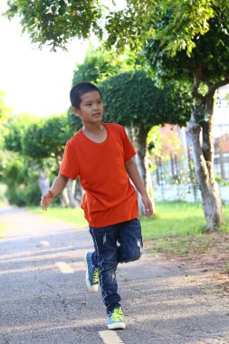 Genç bir çocuk parlak turuncu bir gömlek ve yeşil ayakkabılar giyerek bir patika boyunca neşeyle koşar. Sahne, açık havada huzurlu bir ortamda enerji ve mutluluk yakalar..