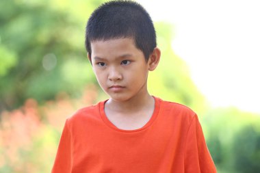 Parlak turuncu tişört giyen genç bir çocuk, güzel bir arka planda ciddi bir yüz ifadesi sergiliyor, bir anlık tefekkür görüntüsü yakalıyor..