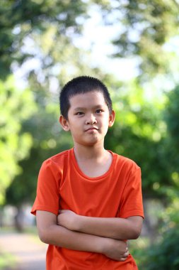 Düşünceli bir çocuk açık havada canlı turuncu bir gömlekle dikiliyor, yemyeşil ile çevrili. Görüntü bir sakinlik ve içgözlem anını yakalar..