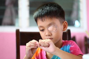 Göz bandı takan genç bir çocuk evde elle yapılan bir aktiviteye odaklanmış, canlı bir iç mekan ortamında masumiyet ve direnç sergiliyor..