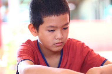 Kırmızı gömlekli genç bir çocuk düşünceli bir şekilde bir aktiviteye odaklanmış oturuyor. Yüzündeki ifade ciddiyet ve masumiyetin karışımını yansıtıyor, çocukluğun özünü yakalıyor..