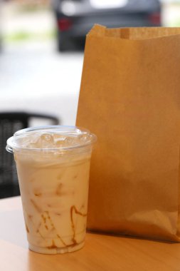 Kremalı ve karamel kıvrımlı ferahlatıcı bir buzlu kahve sıradan bir kafe ortamında keyif almak için mükemmel bir anı somutlaştıran basit bir kese kağıdının yanında oturur..