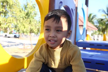 Neşeli bir çocuk, neşeli bir oyun parkında oturur, güneşin ve renkli oyun yapılarının tadını çıkarırken, sıcak bir süveter giyer, tasasız çocukluğun özünü somutlaştırır..