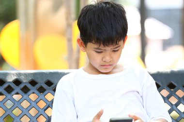 Genç bir çocuk parkta bir bankta oturuyor, akıllı telefonuyla meşgul, huzurlu bir ortamda çocukluk ve dijital teknolojinin kesişimini yansıtıyor..