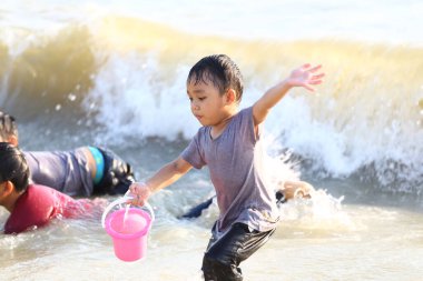 Küçük bir çocuk kumsalda güneşli bir günün tadını çıkarıyor, pembe bir kovayla oynuyor ve suya su sıçrıyor, yaz ve çocukluğun kaygısız ruhunu somutlaştırıyor..