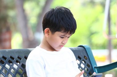 Genç bir çocuk akıllı telefonuyla uğraşırken parkta sessizce oturur, çocukluk merakının ve modern teknolojinin kesişimini canlı bir açık hava ortamında resmeder..