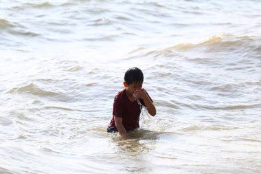 Neşeli genç bir çocuk, sahilde sığ sularda yürürken, yaz eğlencesinin ve maceranın ruhunu yakalar..