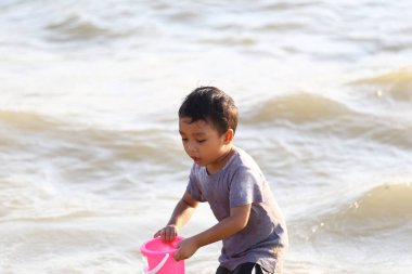Genç bir çocuk kumsalda güneşli bir günün tadını çıkarıyor, suda pembe bir kovayla oynuyor. Sahne çocukluk sevincini ve doğanın güzelliğini yakalar..