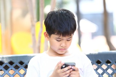 Parktaki bir bankta oturan genç bir çocuk, akıllı telefonuna derinden odaklanmış, dijital dünyada bir anlık konsantrasyon ve bağlılığı somutlaştırıyor..