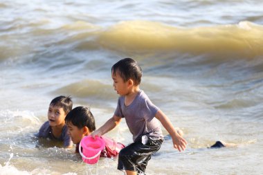 Üç neşeli çocuk plaj aktivitelerine katılıyor, suda oynuyor ve kovalarla eğleniyorlar. Sahne, okyanusta yazın ve çocukluk mutluluğunun özünü yakalar..