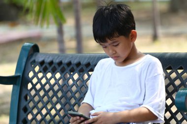 Parktaki bankta oturan genç bir çocuk, akıllı telefonuna dalmış, modern çocukluğu ve günümüz dünyasında çocukların teknolojiyle olan derin bağlantılarını yansıtıyor..