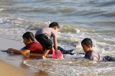 Güneşli bir plaj gününde üç genç, su ve kumla oynayarak huzurlu bir sahil ortamında neşeli anılar yaratıyor..