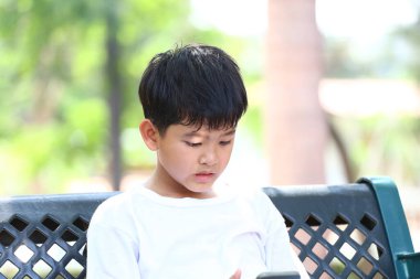 Parktaki bir bankta oturan genç bir çocuk, akıllı telefonuyla meşgul, çevresi yeşillik ve güneş ışığıyla çevrili, modern gençliğin doğadaki çatışmasını resmediyor..