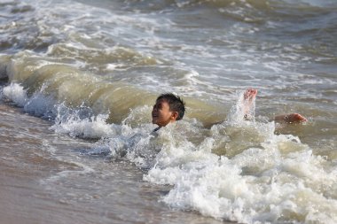 Genç bir çocuk neşeyle okyanus dalgalarında oynuyor, gülüyor ve suya sıçrıyor, güneşli bir günde deniz kenarında çocukluk eğlencesi ve heyecanının özünü yakalıyor..