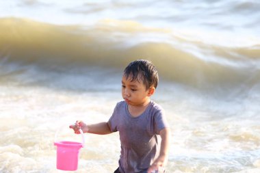 Küçük bir çocuk kumsalda sığ sulara su sıçratıyor, elinde pembe bir kova tutarak, çocukluk ve açık deniz maceralarının neşesini somutlaştırıyor..